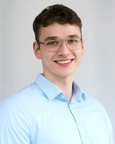 Porträt von David Sättler: lächelnd, mit Brille und hellblauem Hemd vor hellgrauem Hintergrund.